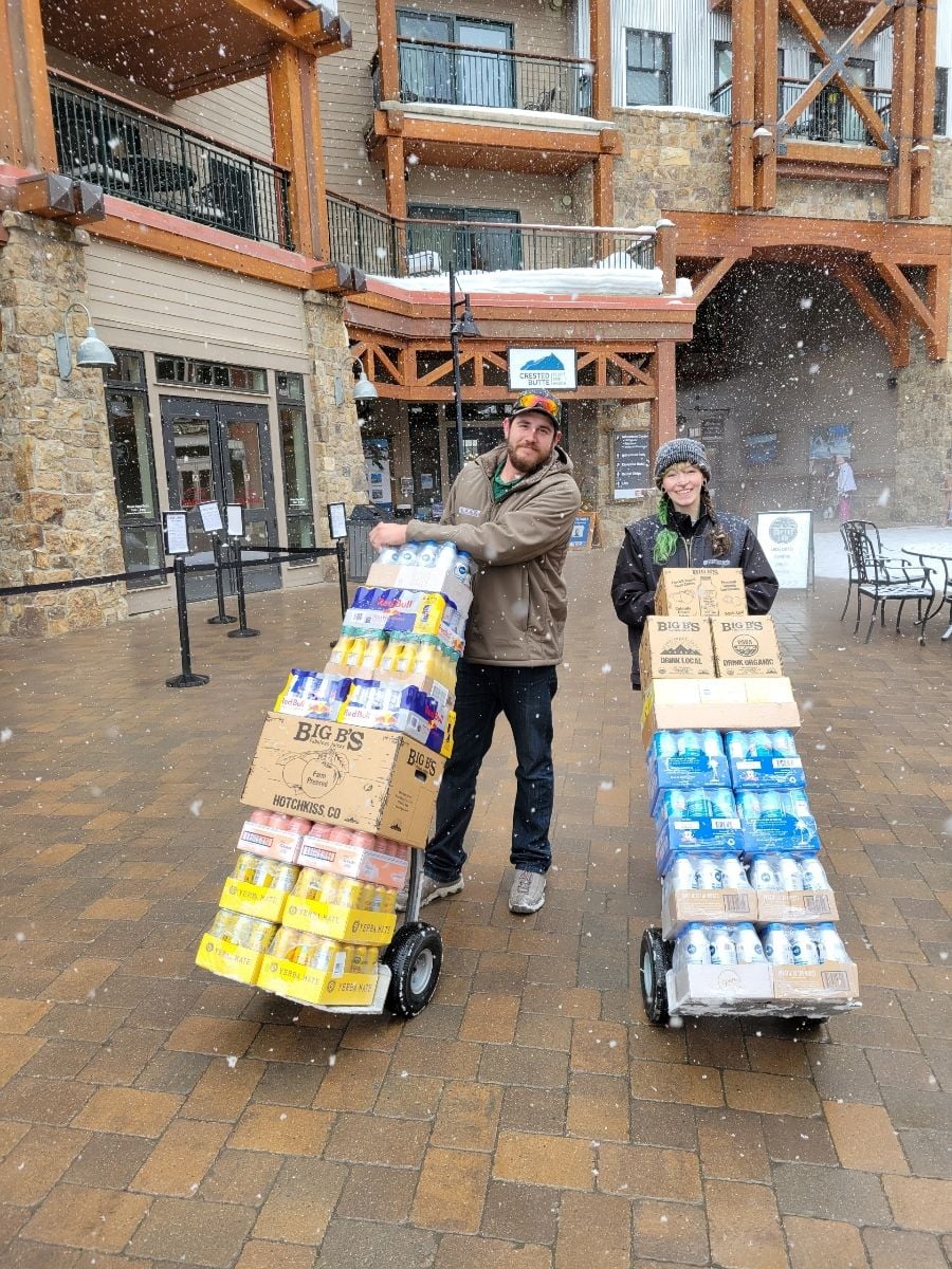 Two Innermountain Distributing team members smiling while delivering stacks of beverages on dollies during a snowfall at a mountain resort.