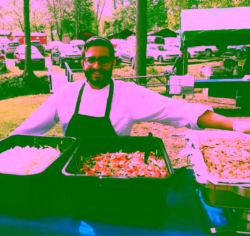 A chef from Deluxe Catering smiling behind a buffet table featuring fresh salads and grilled chicken at an outdoor event.