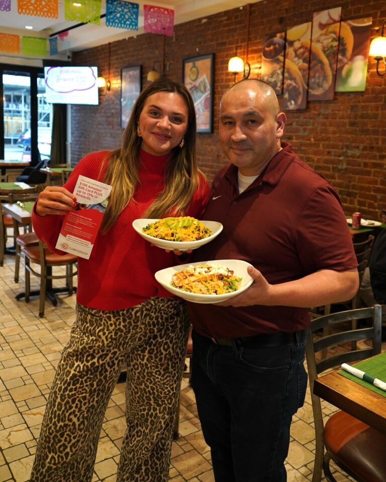 A woman in a red sweater and a man in a maroon polo holding bowls of food and a Caterplace rewards flyer.