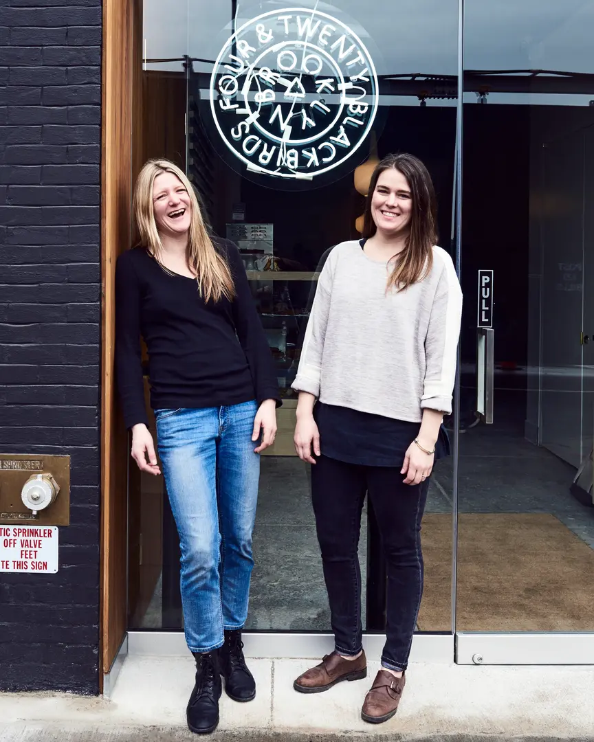 Four & Twenty Blackbirds founders Emily Elsen and Melissa Elsen standing in front of their Brooklyn pie shop's glass door and neon logo.